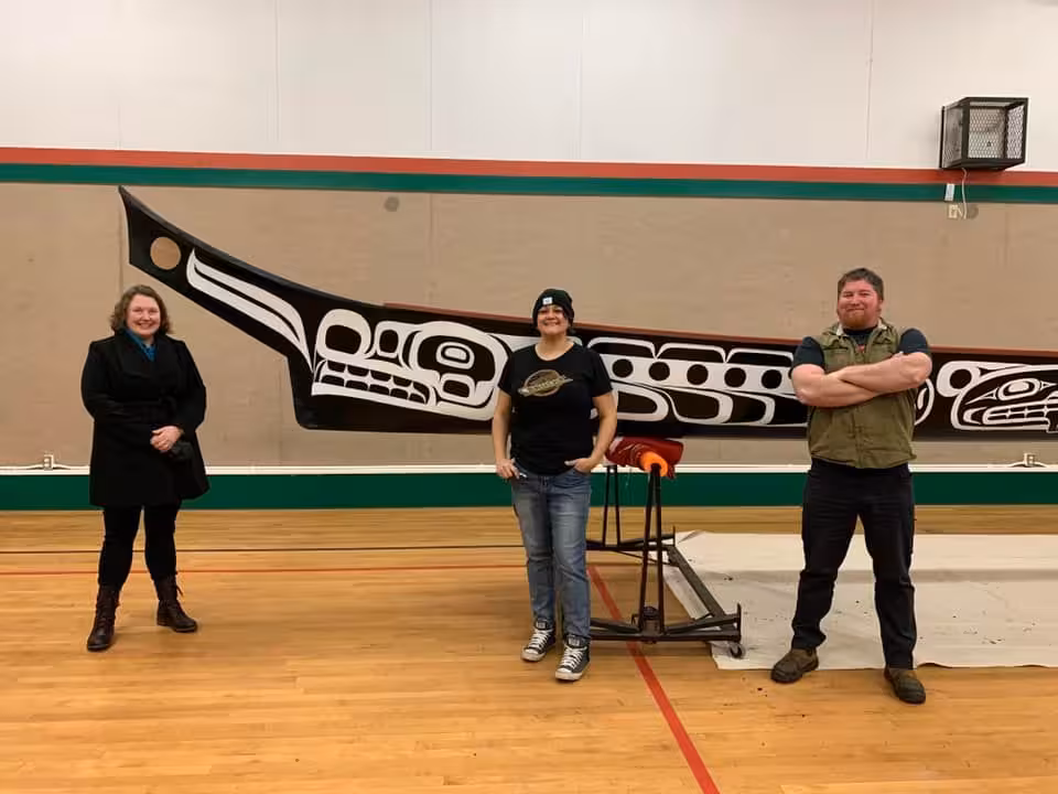 Three K’ómoks First Nation members standing in front of a carved Indigenous canoe in a gymnasium