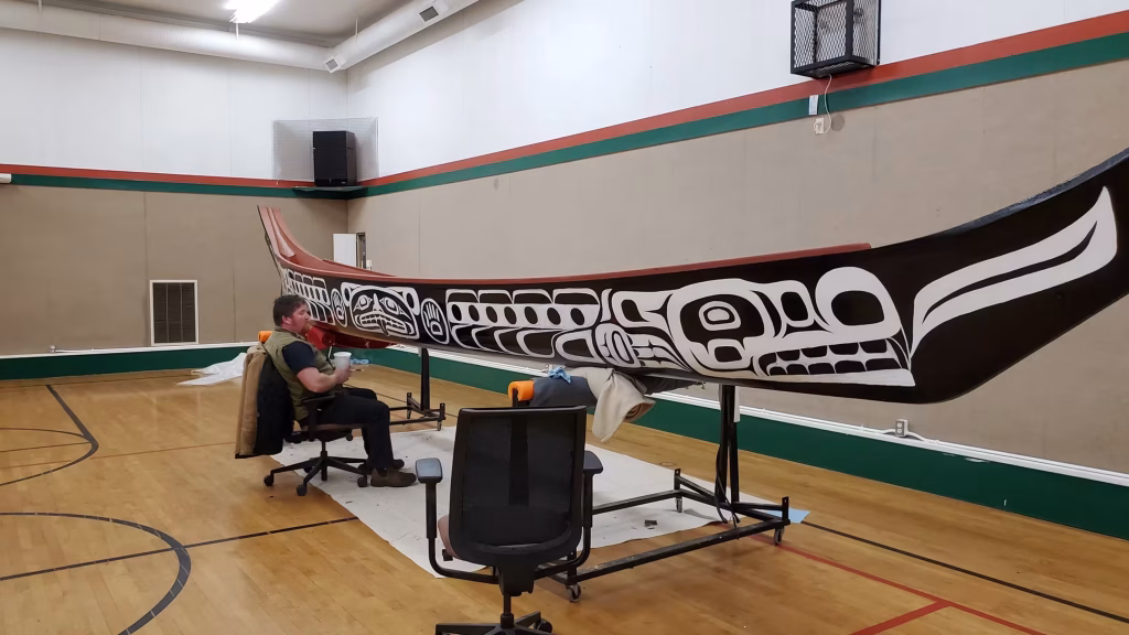 A man painting details on a large K’ómoks First Nation canoe in a gymnasium.