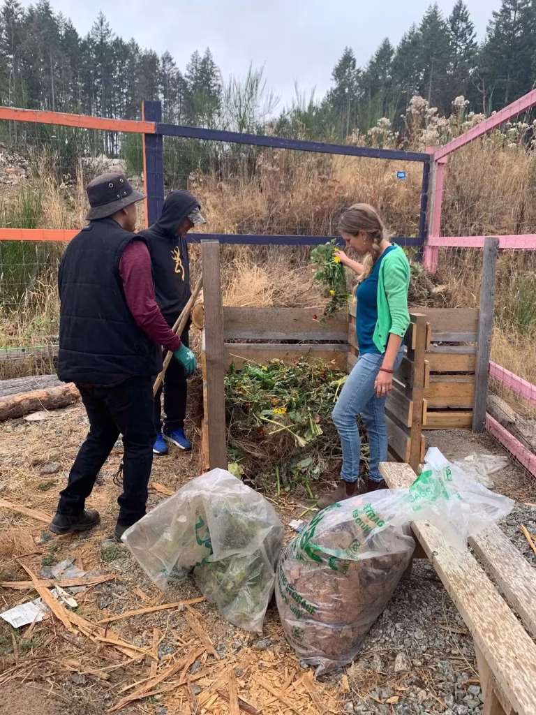 A woman in a green cardigan stands by a wooden compost bin, holding a handful of green plant waste over the pile