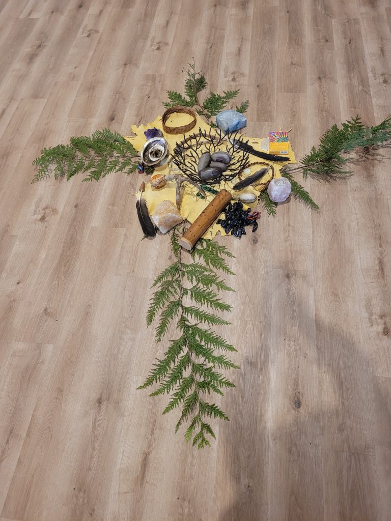 A cross shape is made with natural items including feathers, stones, crystals, and greenery on a wooden floor.