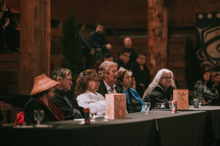 A diverse group of people sit at a table in a dimly lit room with wooden beams, listening attentively.