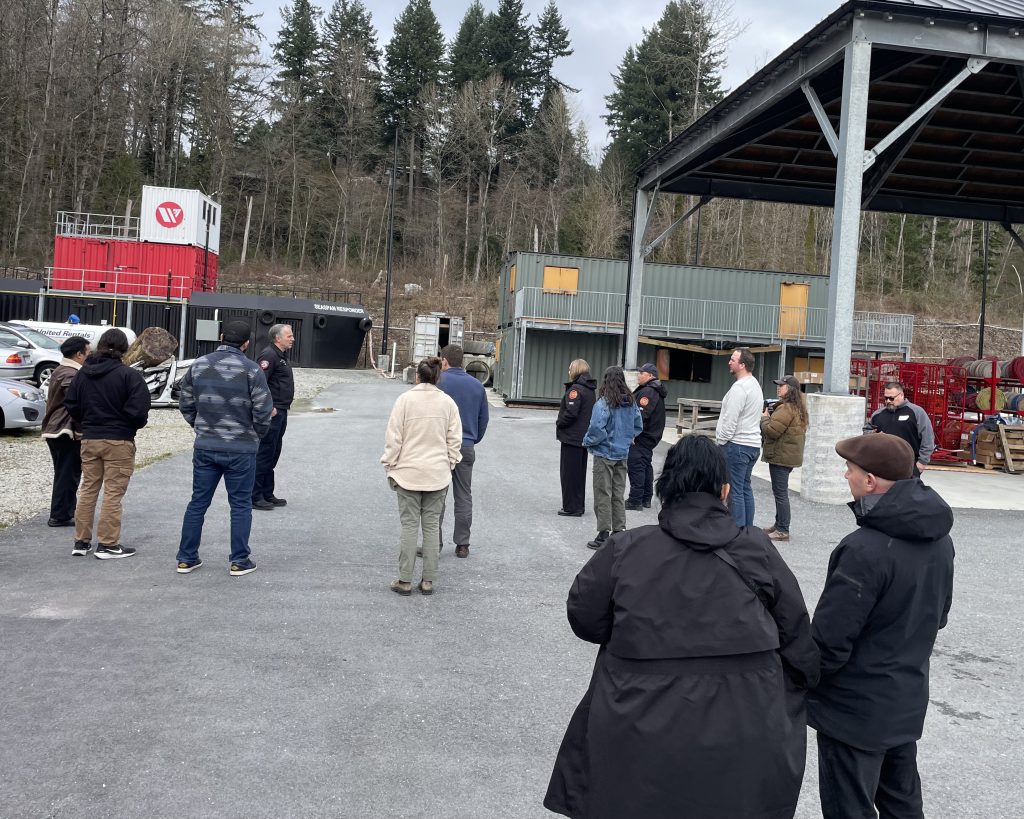 A group of people are gathered outdoors near shipping containers and a covered structure, possibly for a tour or training session.