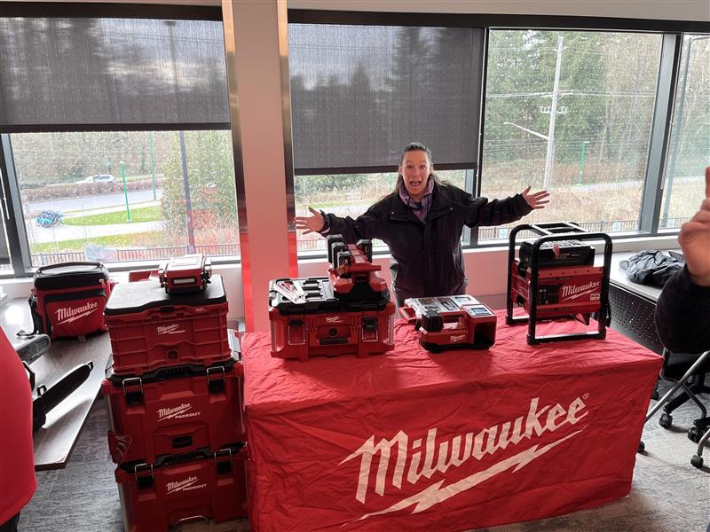 Woman with arms outstretched stands behind a table displaying red Milwaukee toolboxes and equipment.