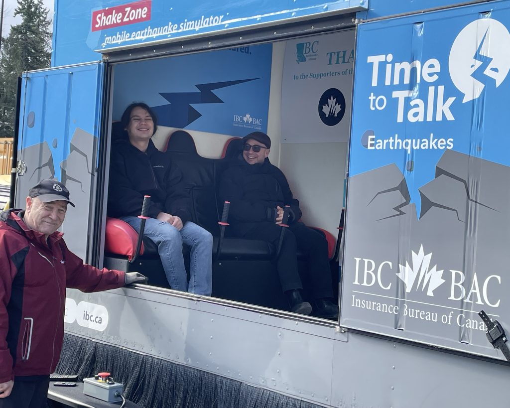 Three people stand in front of and inside a blue mobile earthquake simulator called "Shake Zone.