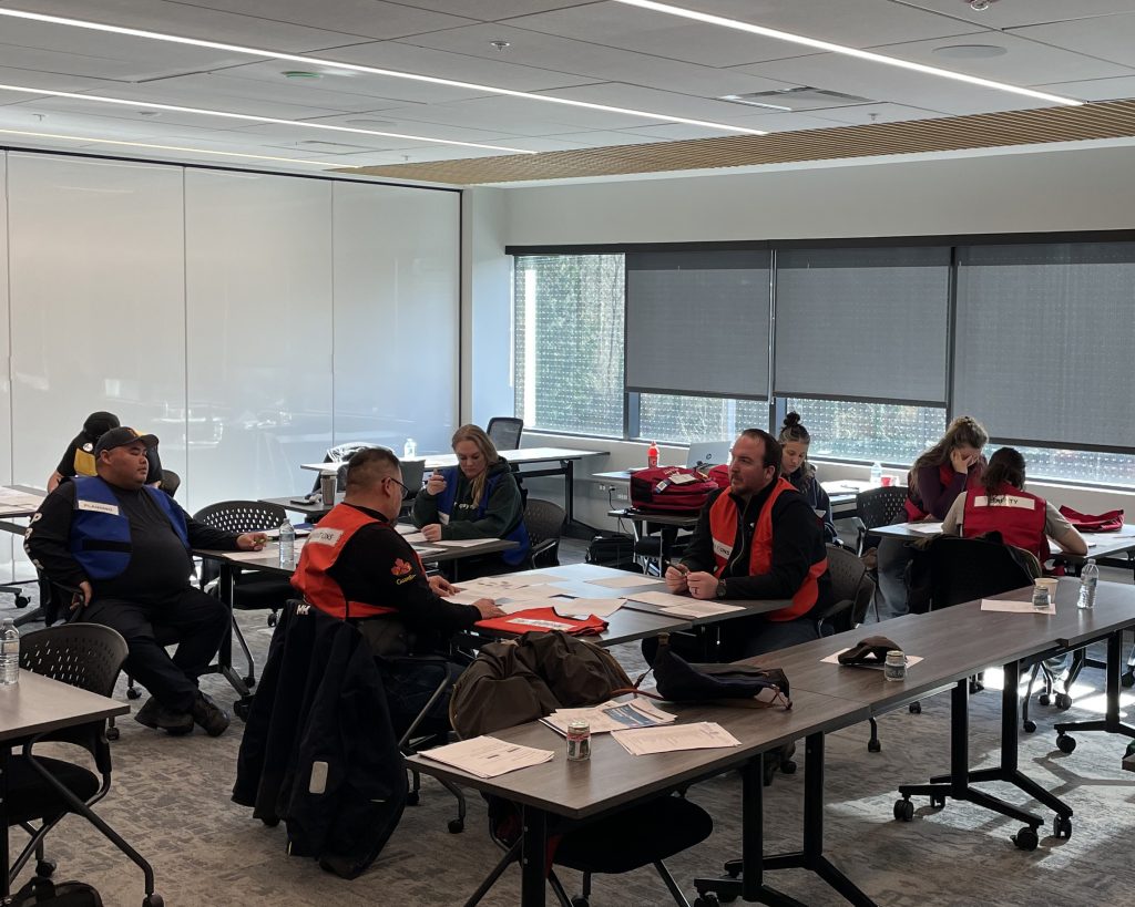 People in a classroom setting wear safety vests and discuss documents at tables.