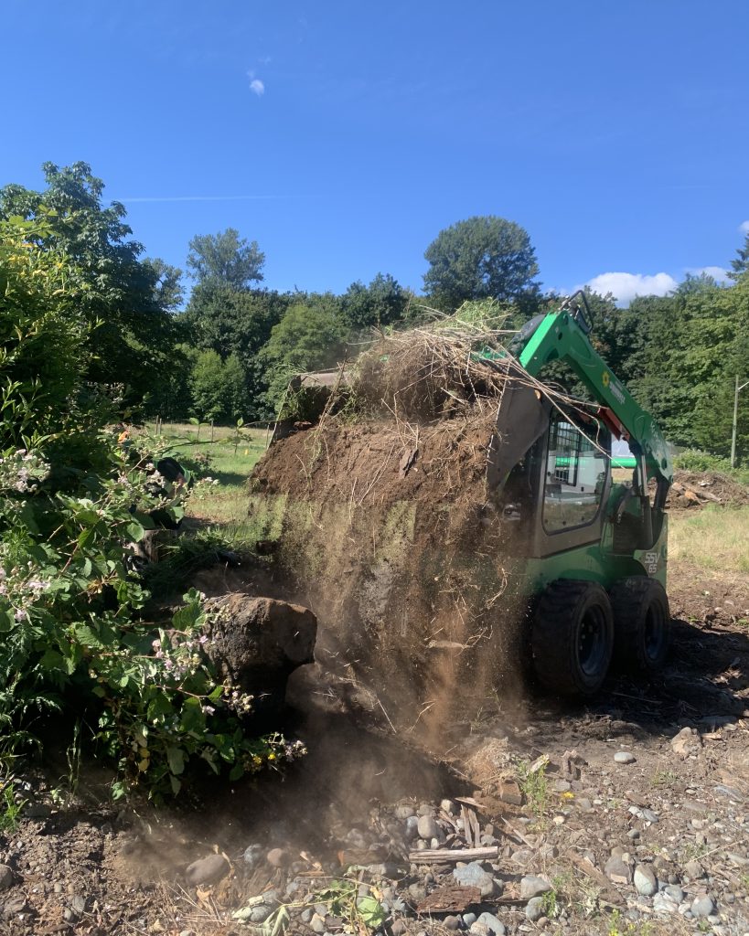 Green skidsteer loader moving a pile of dirt and vegetation on a sunny day.