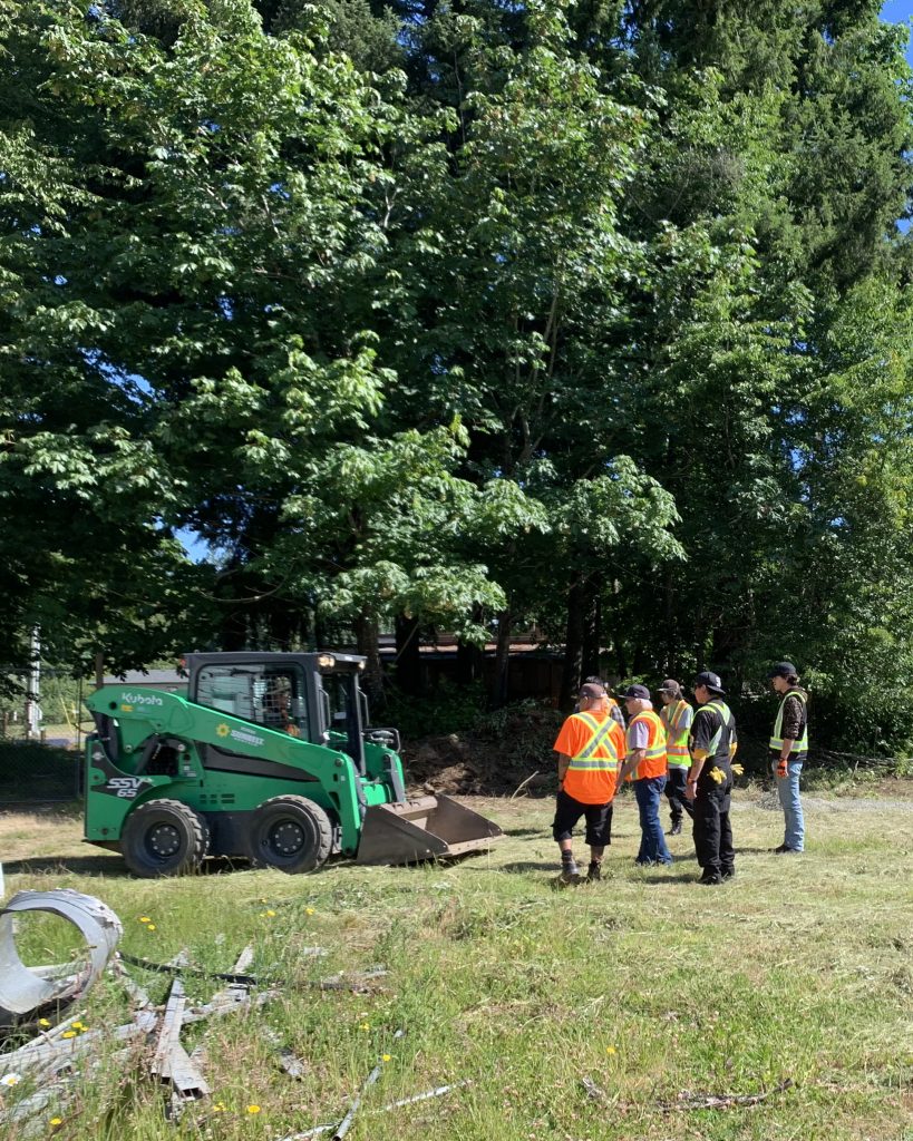 A group of men wearing high-visibility vests stand in a grassy field near a green Kubota skid steer loader.