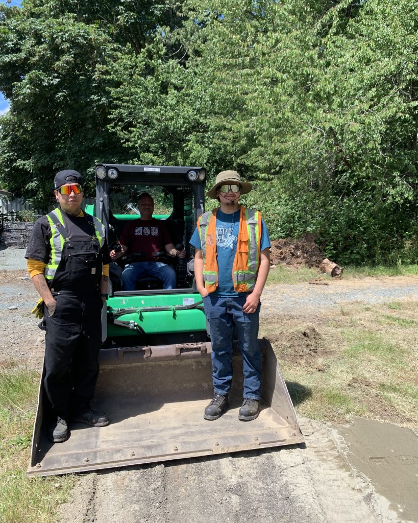 Three workers stand in front of a skid steer loader outdoors on a sunny day.