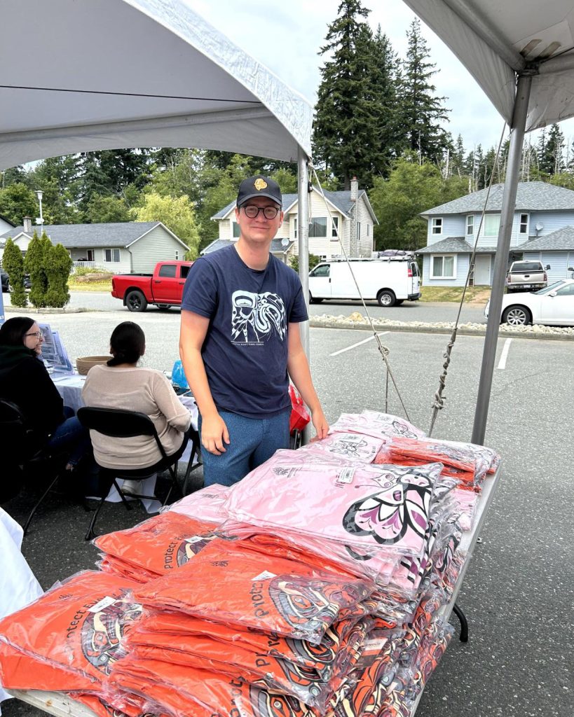 Man in a dark blue t-shirt with graphic print and a baseball cap stands behind a table of stacked, packaged t-shirts.