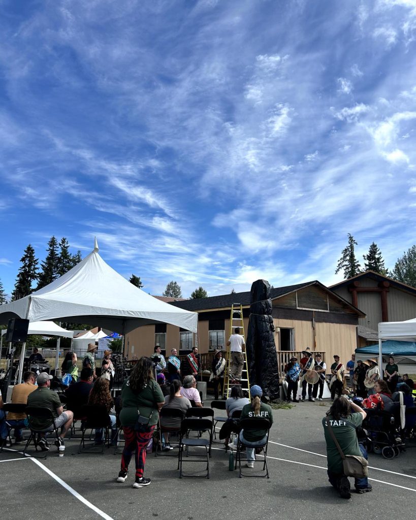 Outdoor gathering with a crowd watching a performance by people in traditional regalia near a large totem pole.
