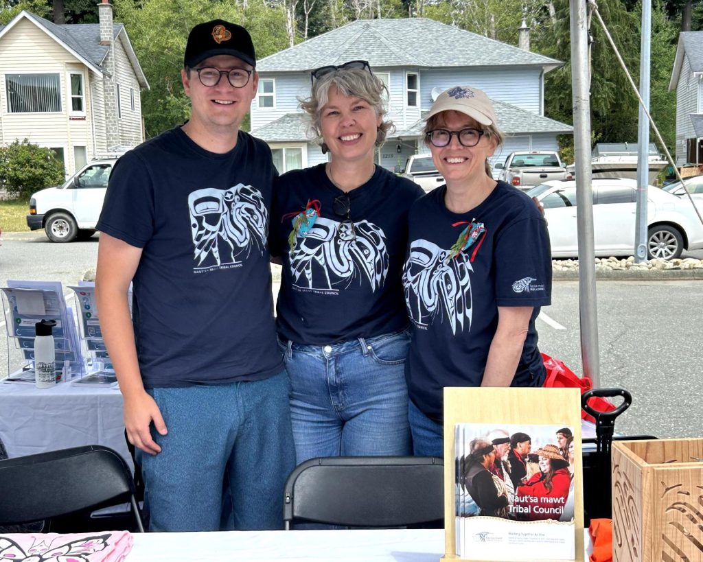 Three people smiling at a table outdoors, wearing matching t-shirts with Indigenous art.