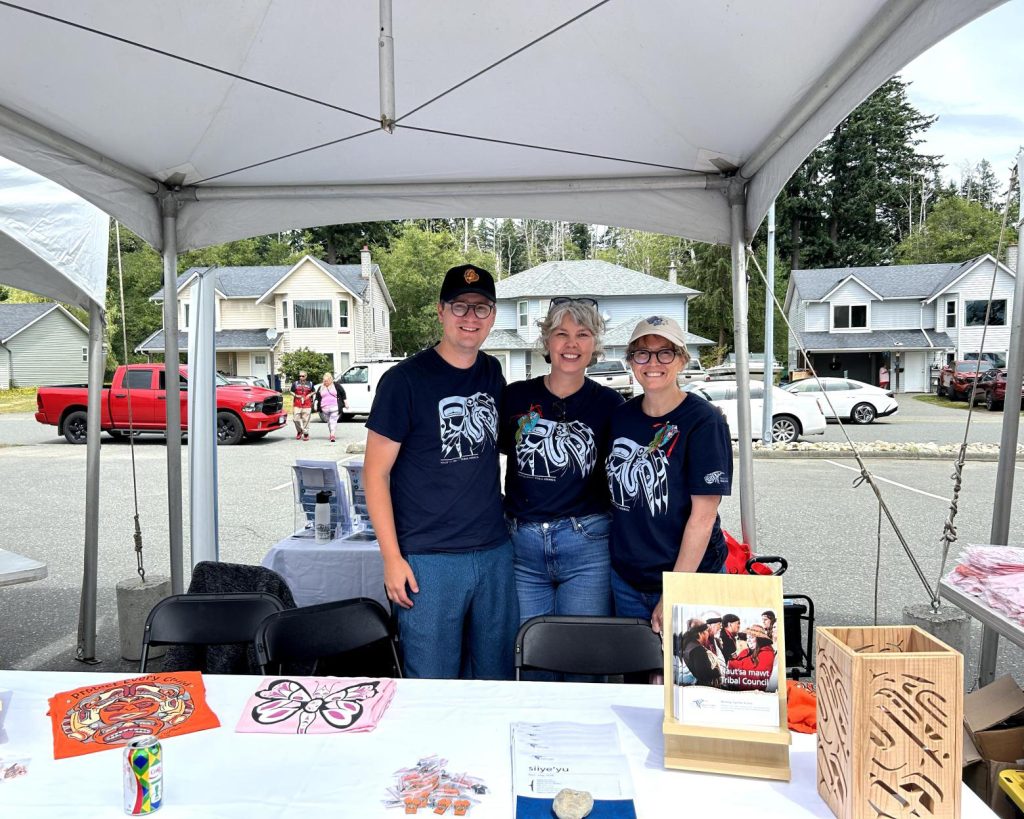 Three people stand under a tent at an outdoor event, smiling at the camera.
