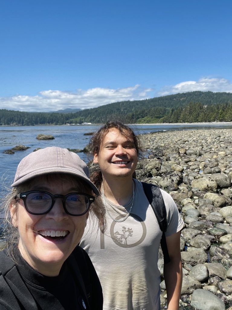 Two people smile for a selfie on a rocky beach with a forested coastline and cloudy sky in the background.