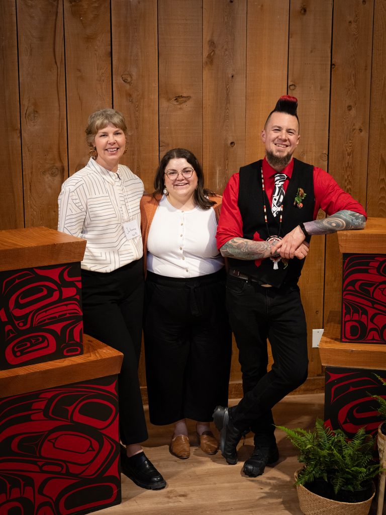 Three people pose in front of a wooden wall, with Indigenous art boxes on either side.