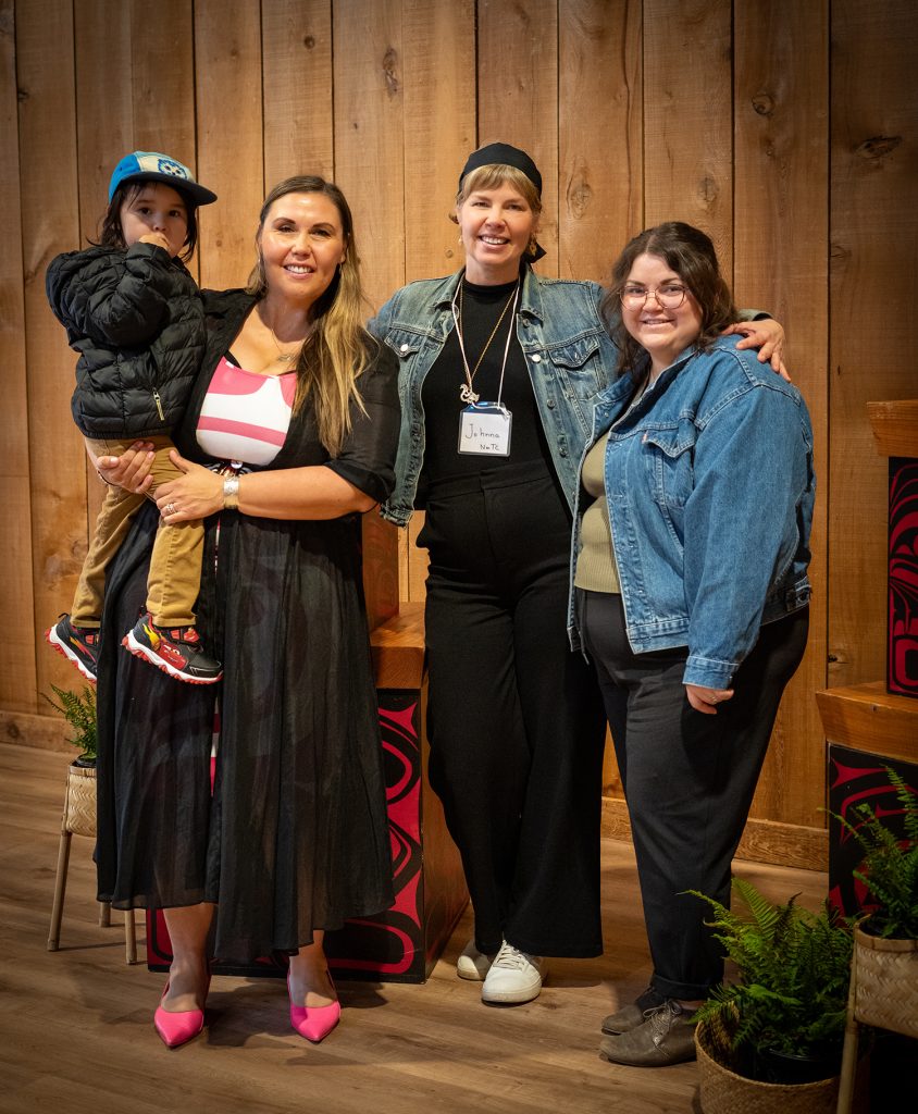 A woman holds a young boy while standing with two other women in front of a wooden wall.