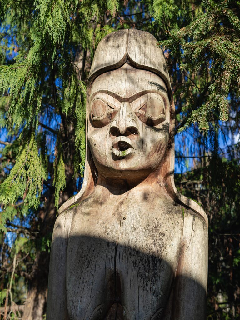 Close-up of a weathered wooden totem pole with a carved face, set against green trees and blue sky.