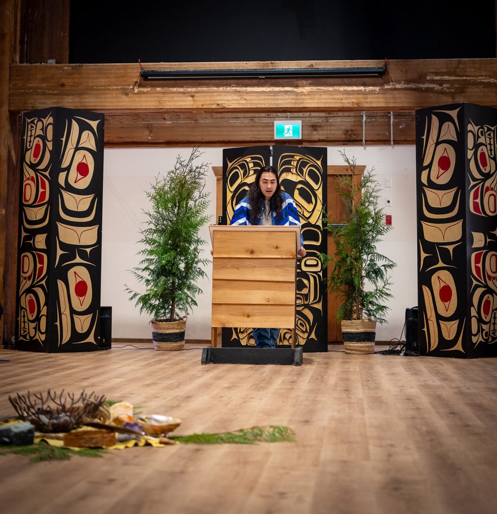 A person stands at a wooden podium in front of a large, intricately designed backdrop, flanked by two tall, decorated columns and potted plants.