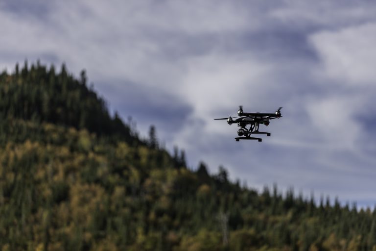 A black drone with a camera is flying in the sky above a tree-covered hill.